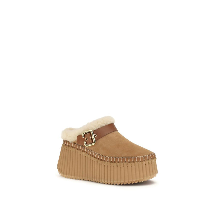 Nama Wedge MulesThe Nama Wedge Mules by Chloé blend casual elegance with artisanal mastery. Crafted from suede calfskin and shearling, they feature a leather strap with the House’s signature Paddington buckle. The lightweight wedge, made from recycled mat