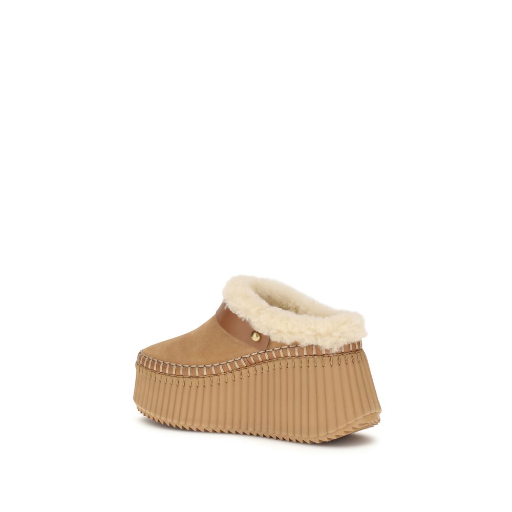 Nama Wedge MulesThe Nama Wedge Mules by Chloé blend casual elegance with artisanal mastery. Crafted from suede calfskin and shearling, they feature a leather strap with the House’s signature Paddington buckle. The lightweight wedge, made from recycled mat