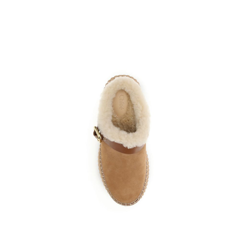 Nama Wedge MulesThe Nama Wedge Mules by Chloé blend casual elegance with artisanal mastery. Crafted from suede calfskin and shearling, they feature a leather strap with the House’s signature Paddington buckle. The lightweight wedge, made from recycled mat